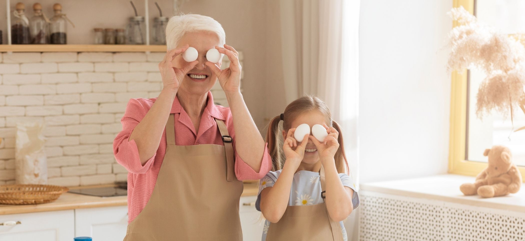 an older woman an a young girl stand in a kitchen holding eggs in front of their eyes