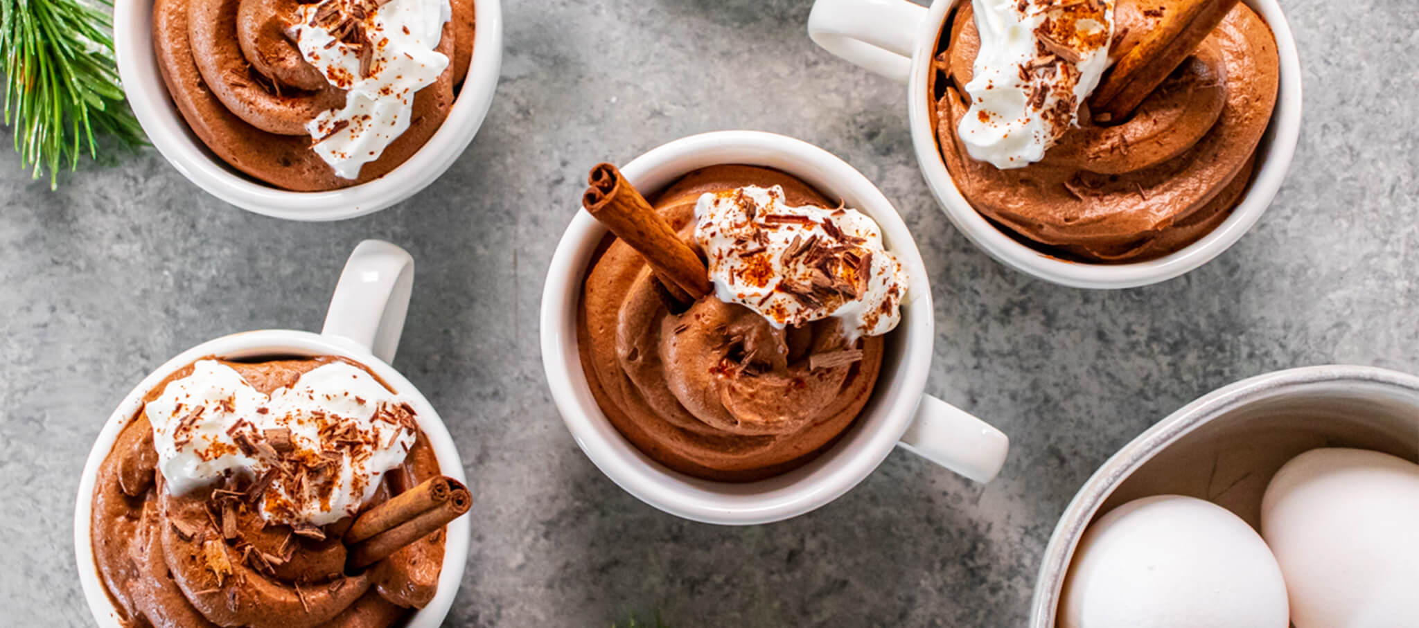 Four mugs of chocolate mousse topped with cream and cinnamon sticks
