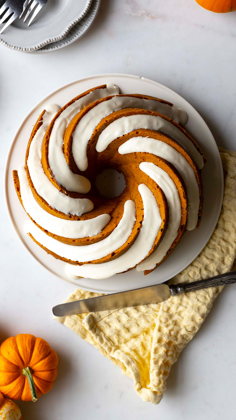 A spiral-shaped pumpkin bundt cake drizzled with white icing, placed on a white plate over a yellow textured cloth, surrounded by small decorative pumpkins and utensils.
