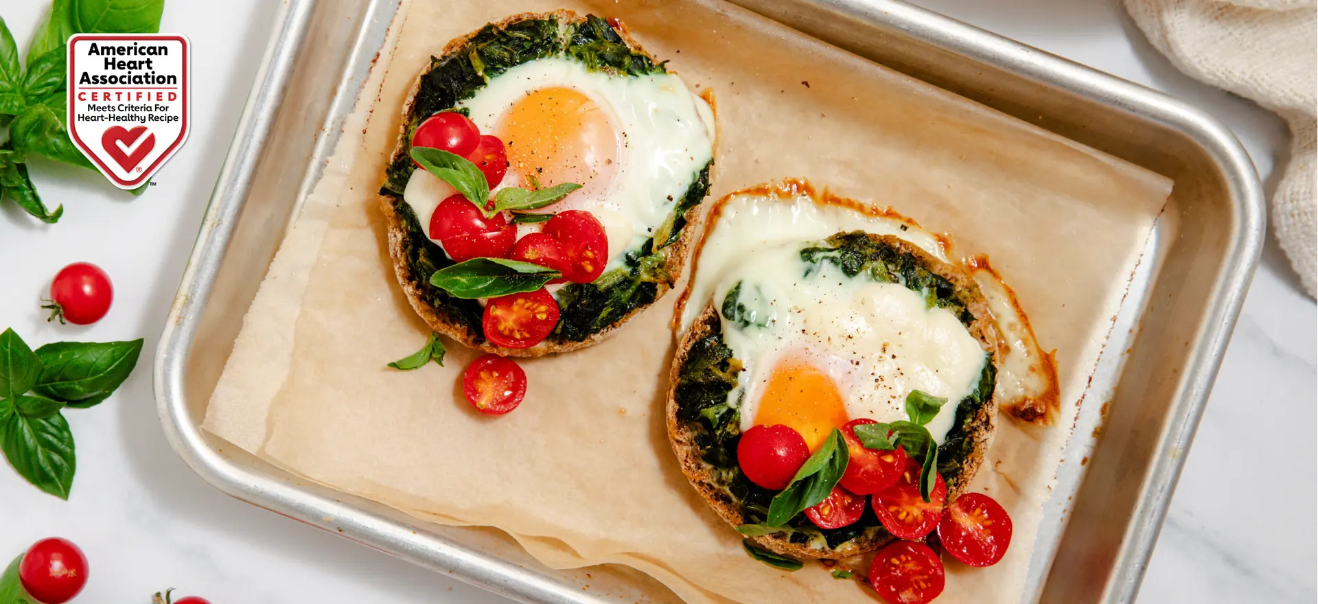 Two mini breakfast pizzas on a baking sheet with spinach, fried eggs, tomatoes, and basil. Includes American Heart Association Certified logo.