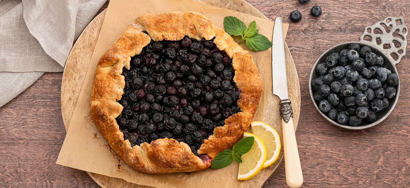 Homemade blueberry galette on parchment with a flaky crust, served beside a bowl of fresh blueberries and lemon slices